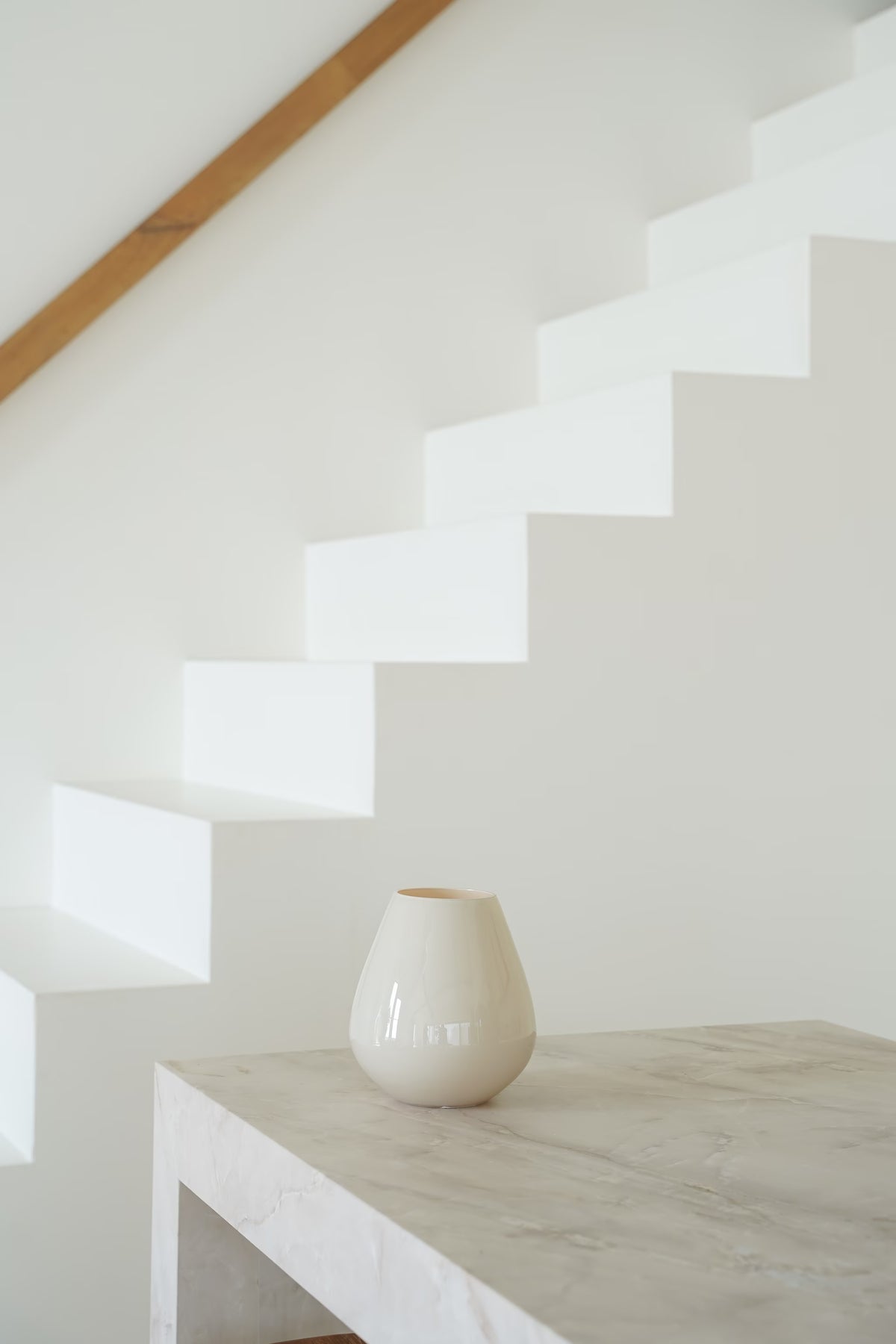 Crèmekleurige Vaas Mora met glanzende afwerking op een lichte stenen sidetable bij een witte trap.