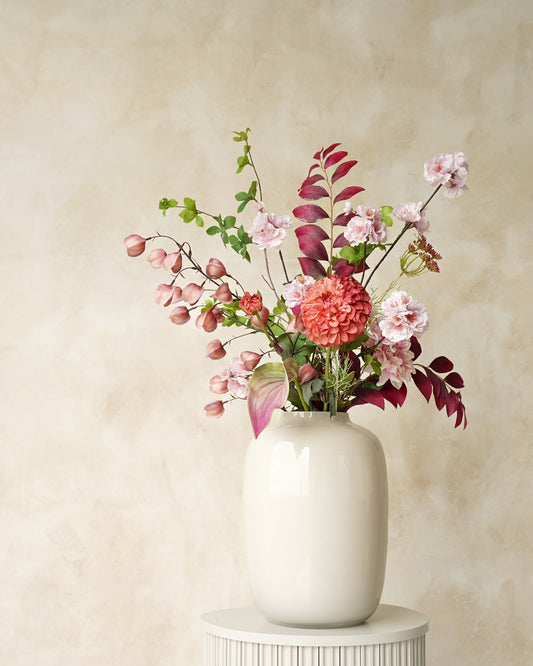 Zijden veldboeket Carmen van Fan of Flowers met zachtroze bloesem, een koraalroze bloem en bordeauxrood blad in witte vaas Crema op een ronde sokkel.