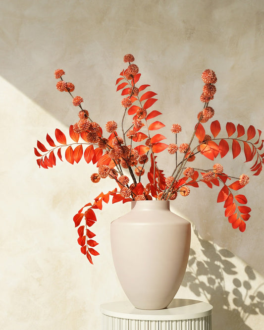 Zijden herfstboeket Fuego van Fan of Flowers met oranje bladtakken en kleine bolbloemen in een lichtbeige vaas op een ronde sokkel.