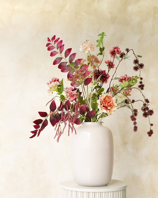 Zijden boeket Lila van Fan of Flowers met bordeauxrode bladeren, roze en crèmekleurige bloemen in witte vaas Crema op een ronde sokkel.