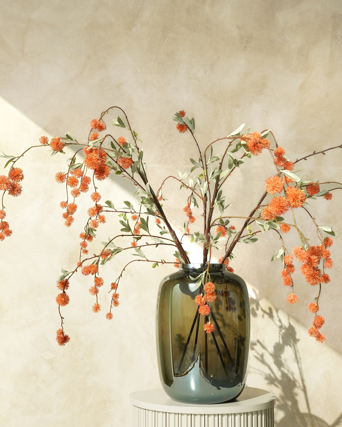 Oranje zijden bolletjestakken van Fan of Flowers met afhangende vertakkingen in een glanzende donkergroene glazen vaas op een ronde sokkel.