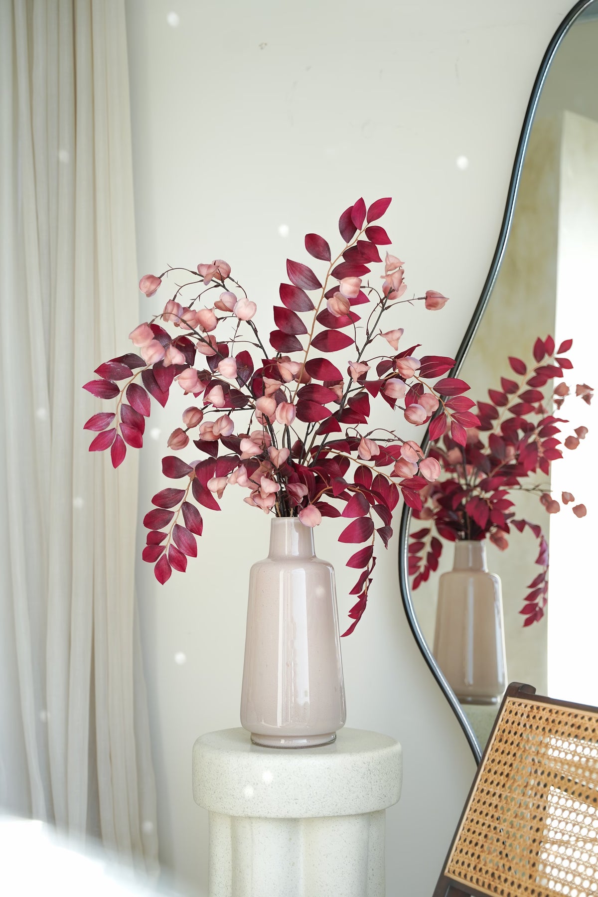 Vaas Lino met bordeauxrode bladertakken en roze lampionvormige decoraties op een lichte sokkel voor een spiegel.