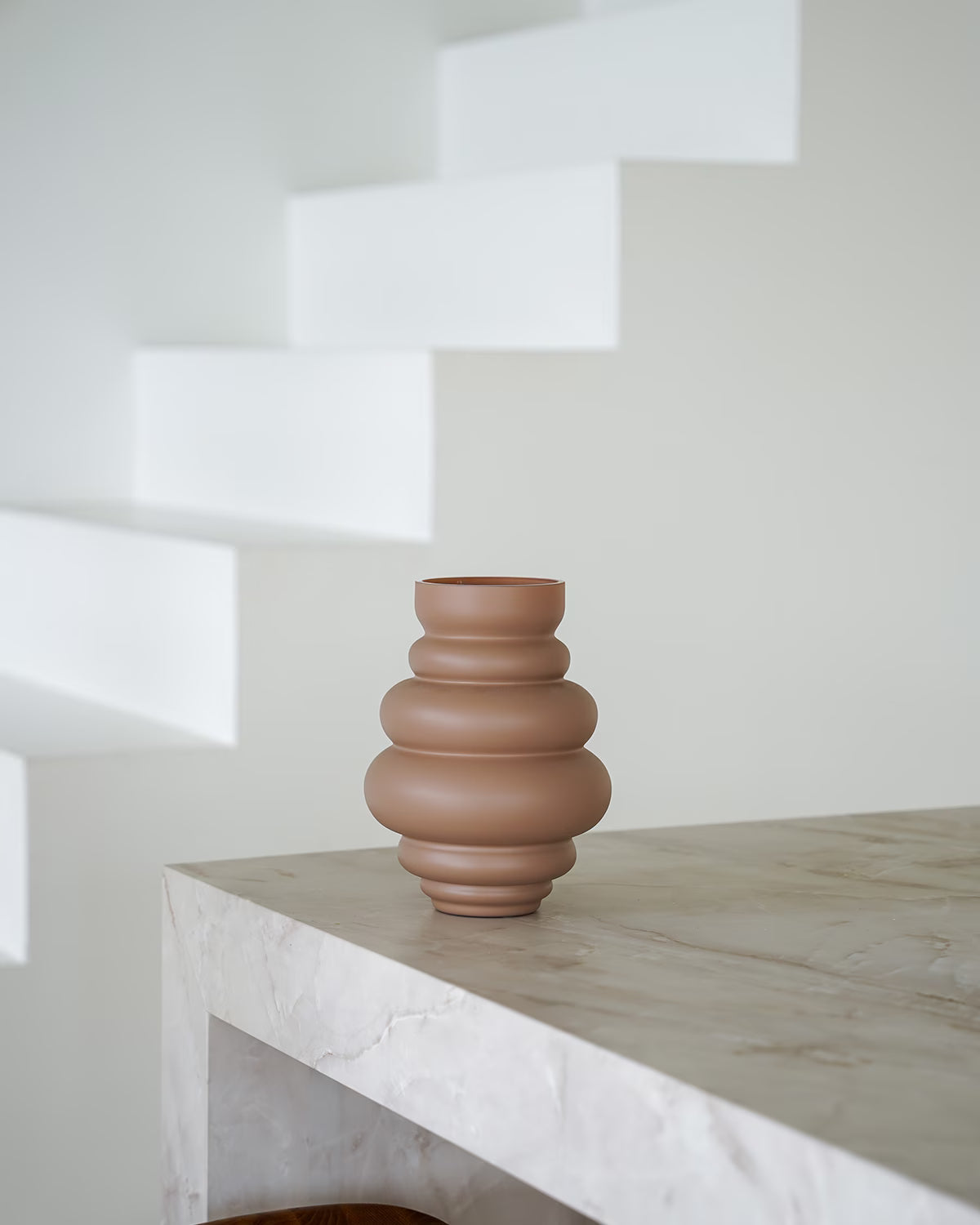 Een lege, matbruine/terracotta Vaas Bomba van Fan of Flowers, maat S, geplaatst op een marmeren bijzettafel tegen een witte achtergrond.