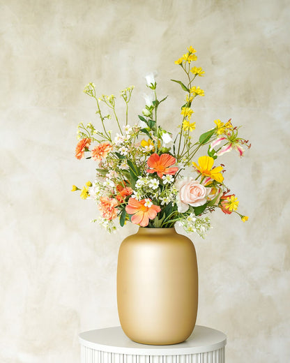 Een gouden Vaas Mateo van Fan of Flowers, gevuld met het vrolijke zijden Boeket Fia in oranje en gele tinten op een witte sokkel.