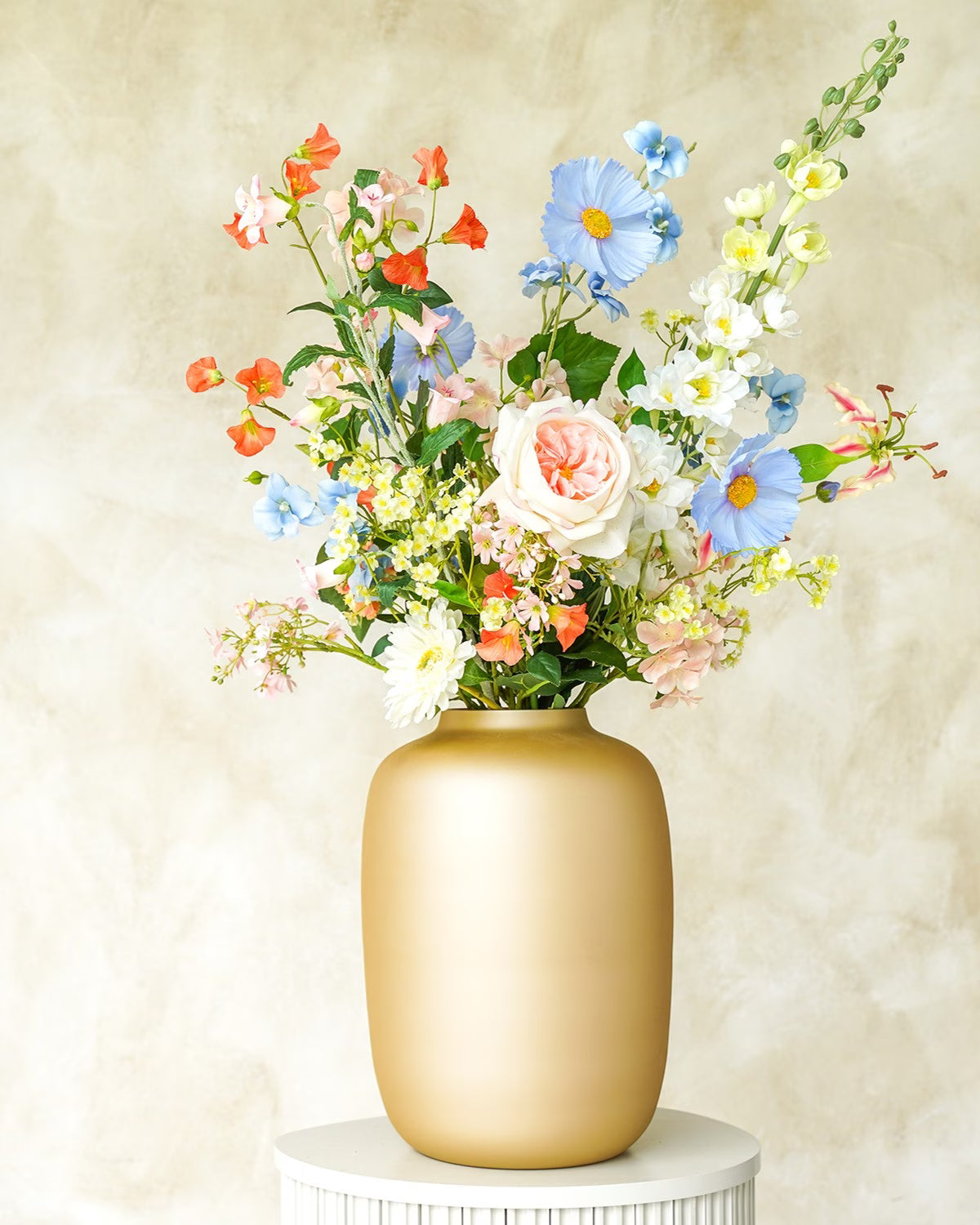 Een gouden Vaas Mateo van Fan of Flowers, gevuld met het kleurrijke zijden Boeket Mari in blauwe, witte en roze tinten op een witte sokkel.