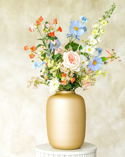 Een gouden Vaas Mateo van Fan of Flowers, gevuld met het kleurrijke zijden Boeket Mari in blauwe, witte en roze tinten op een witte sokkel.