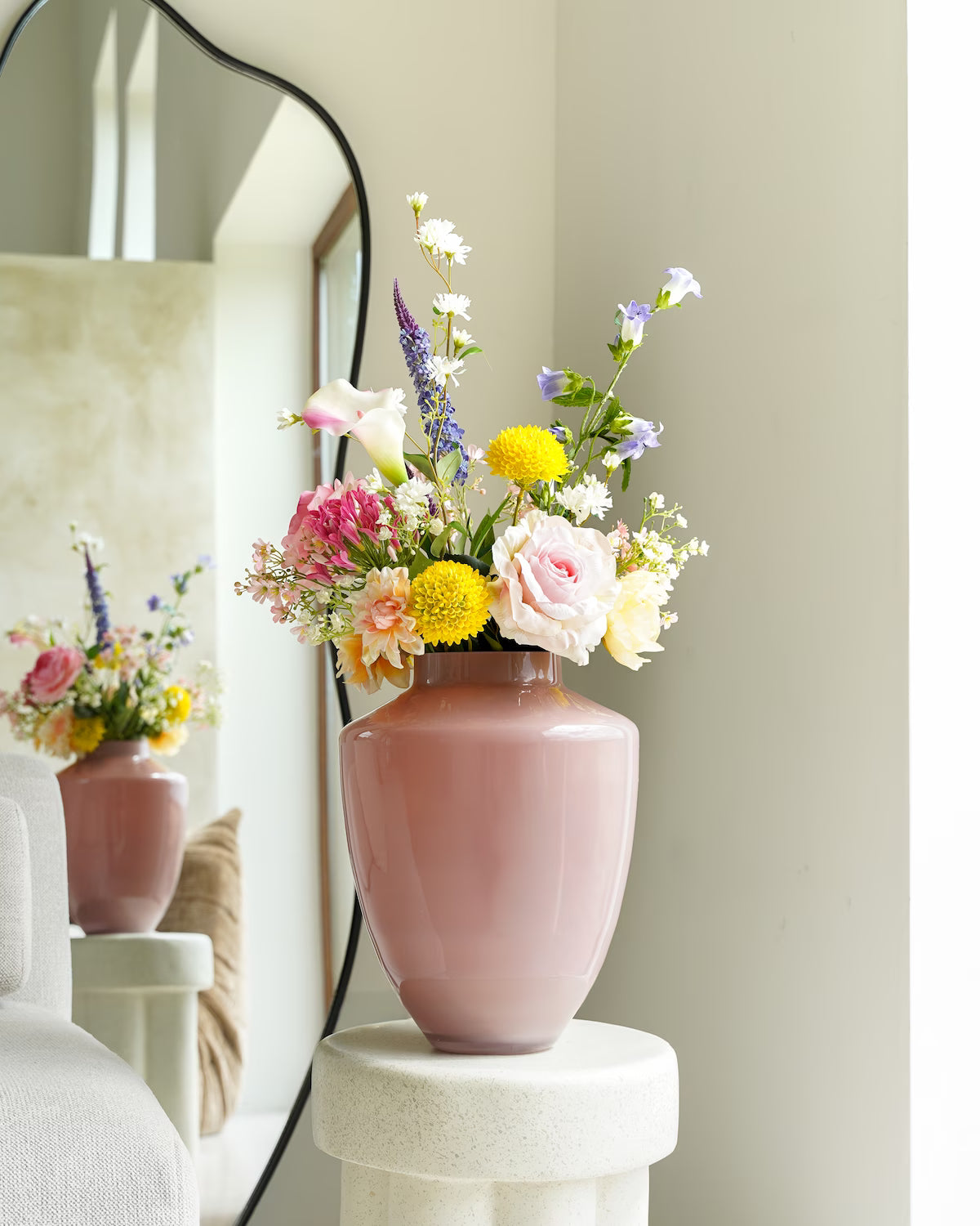 De roze vaas Rosa van Fan of Flowers (maat M) gevuld met een vrolijk, gemengd voorjaarsboeket, staand op een sokkel voor een spiegel.