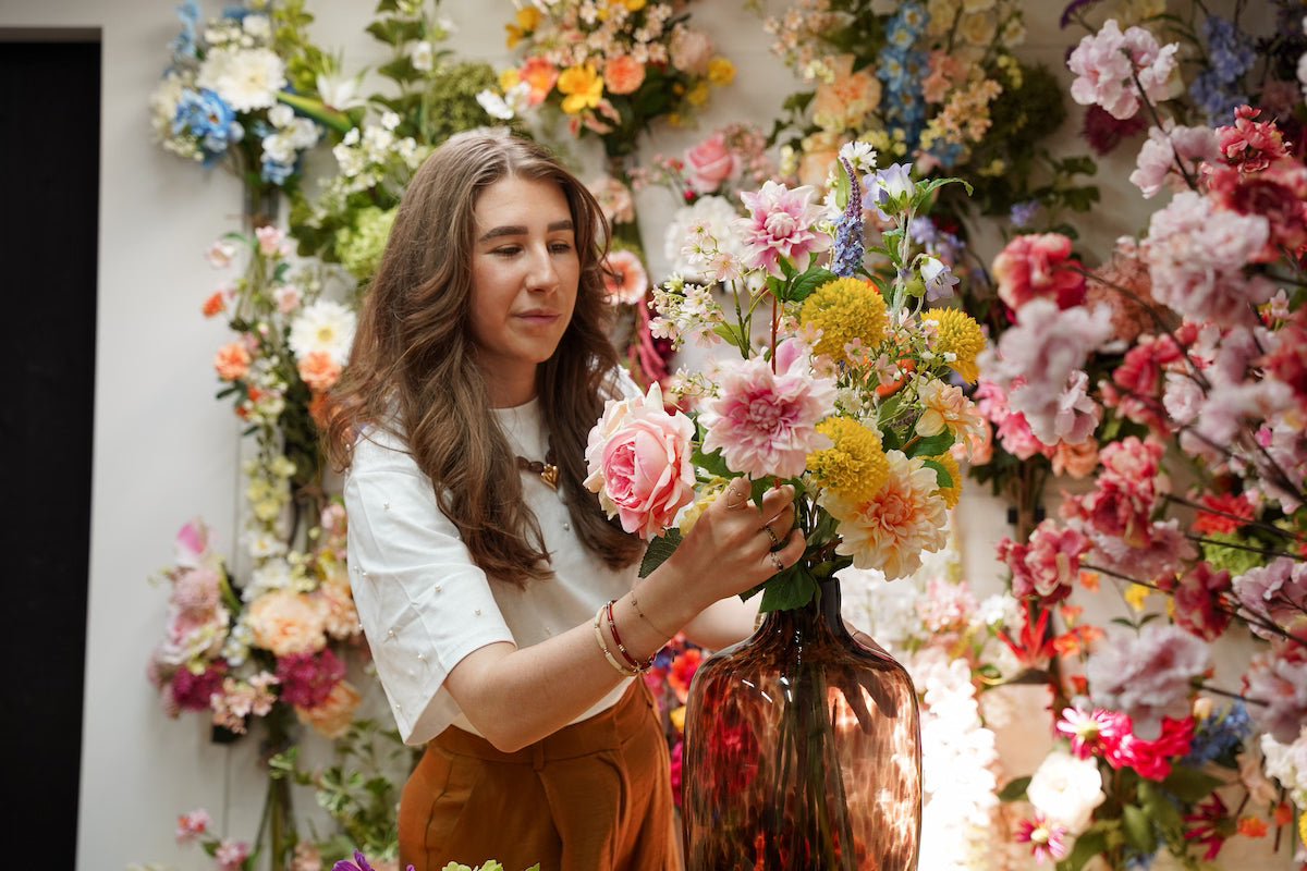 Femke Goossens schikt secuur een vrolijk zijden veldboeket in de bruinglas Vaas Leo in het atelier van Fan of Flowers.