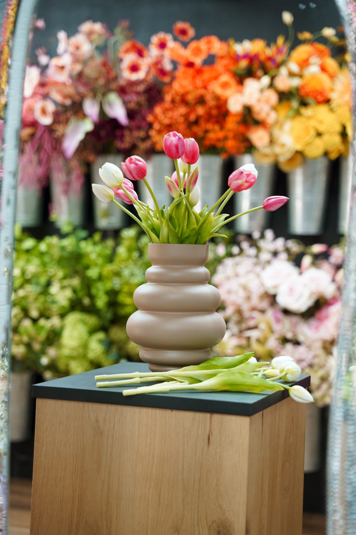 Beige Vaas Bomba met roze en witte zijden tulpen op een houten zuil met bloemen op de achtergrond.
