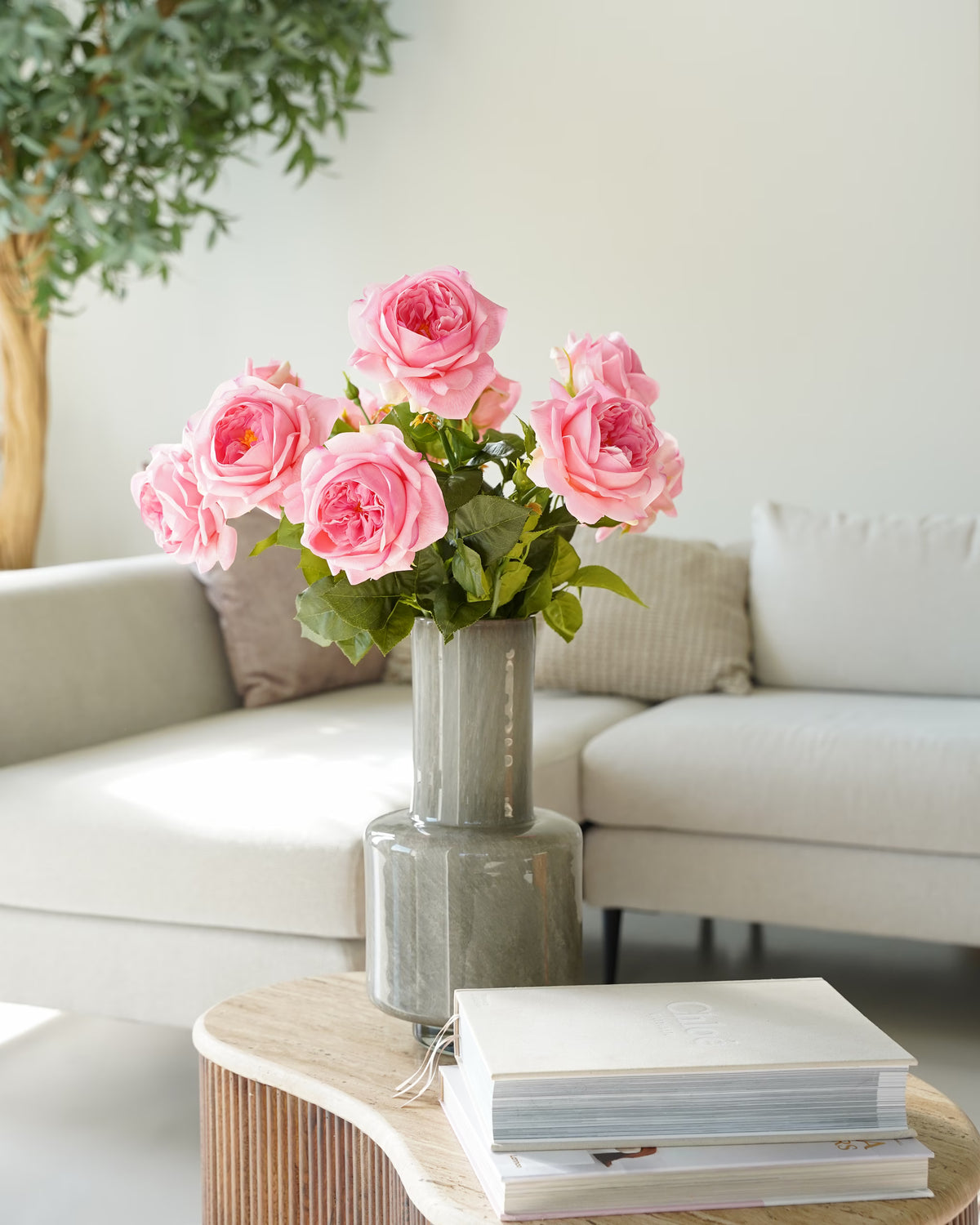 Vaas Humo met roze zijden rozen op een ronde houten salontafel voor een lichte hoekbank.