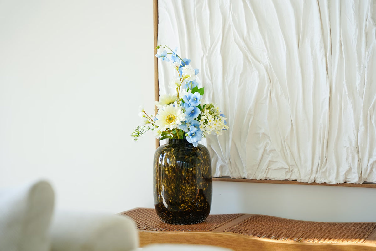 Tijgervaas Leo S van Fan of Flowers met een lichtblauw en wit zijden boeket op een houten bankje voor een wit wandkunstwerk.