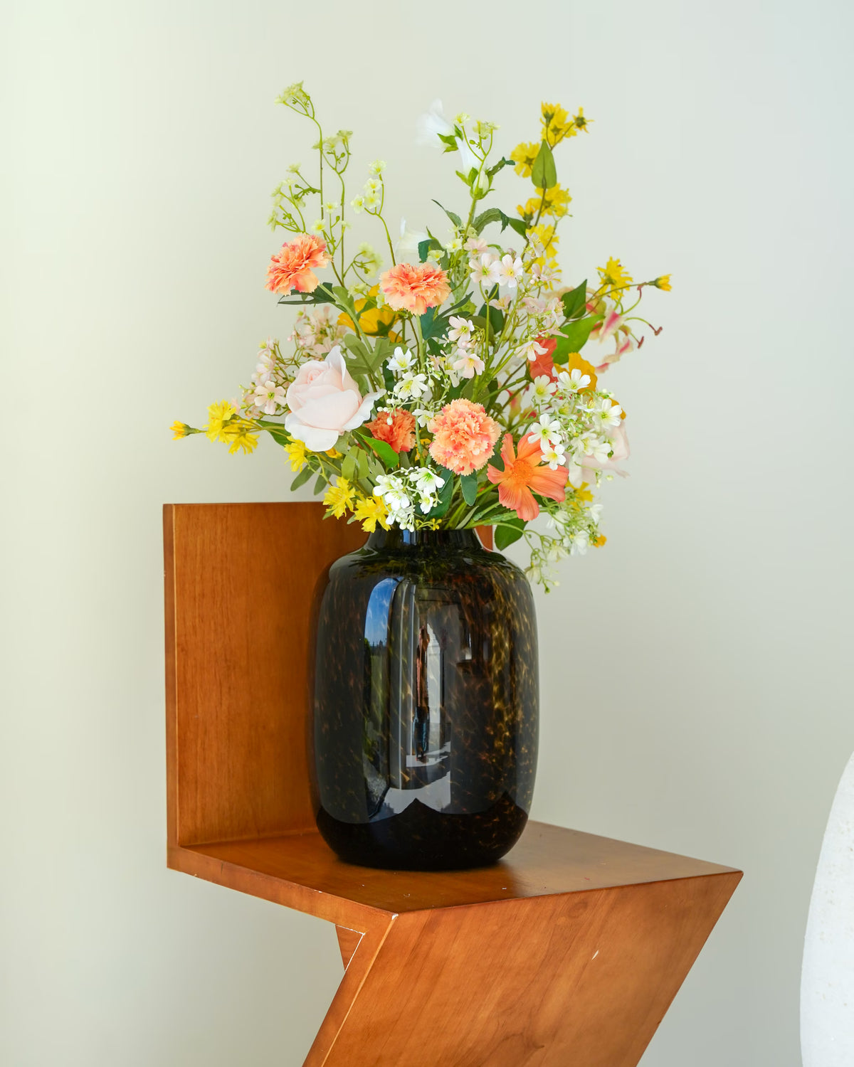 Glazen tijgervaas Leo S van Fan of Flowers met gele, perzikkleurige, zachtroze en witte zijden bloemen op een houten stoel.
