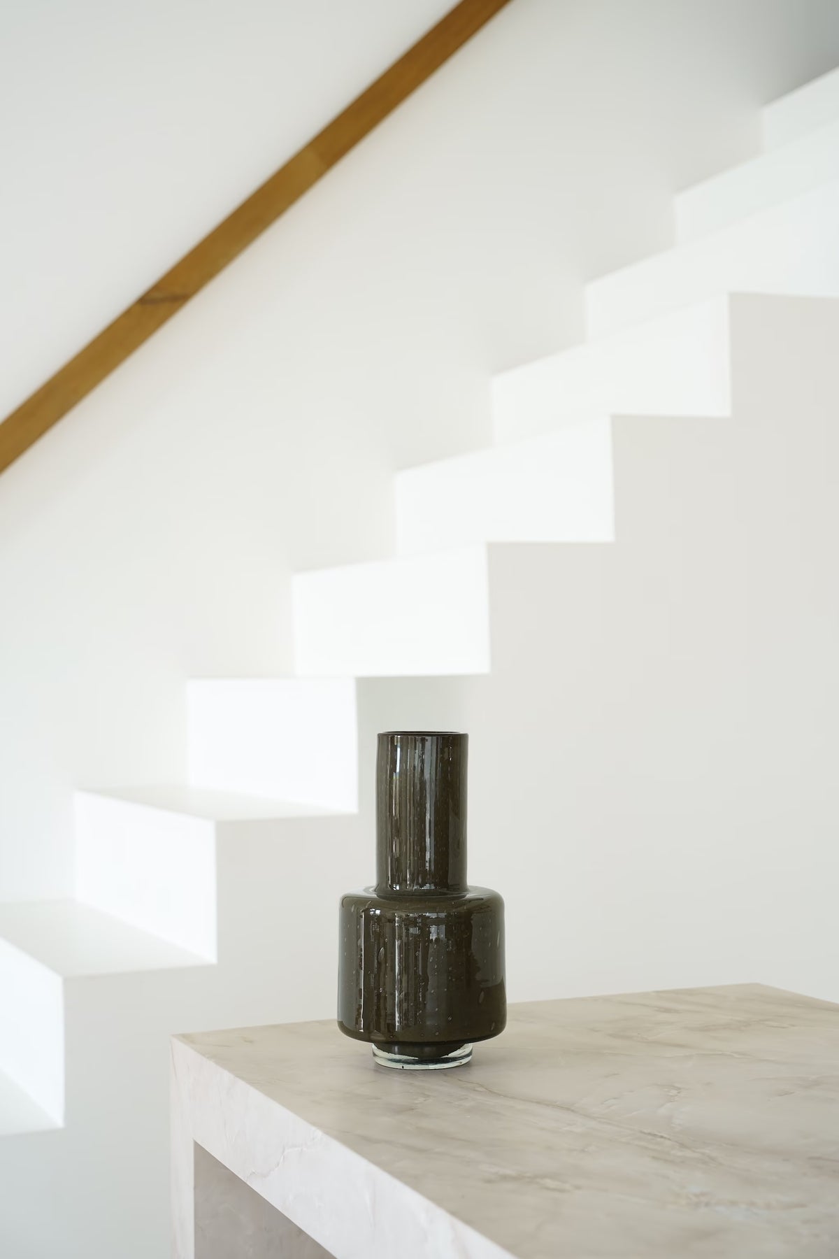 Donkerbruine Vaas Madera met lange hals en heldere glazen voet op een lichte stenen sidetable.