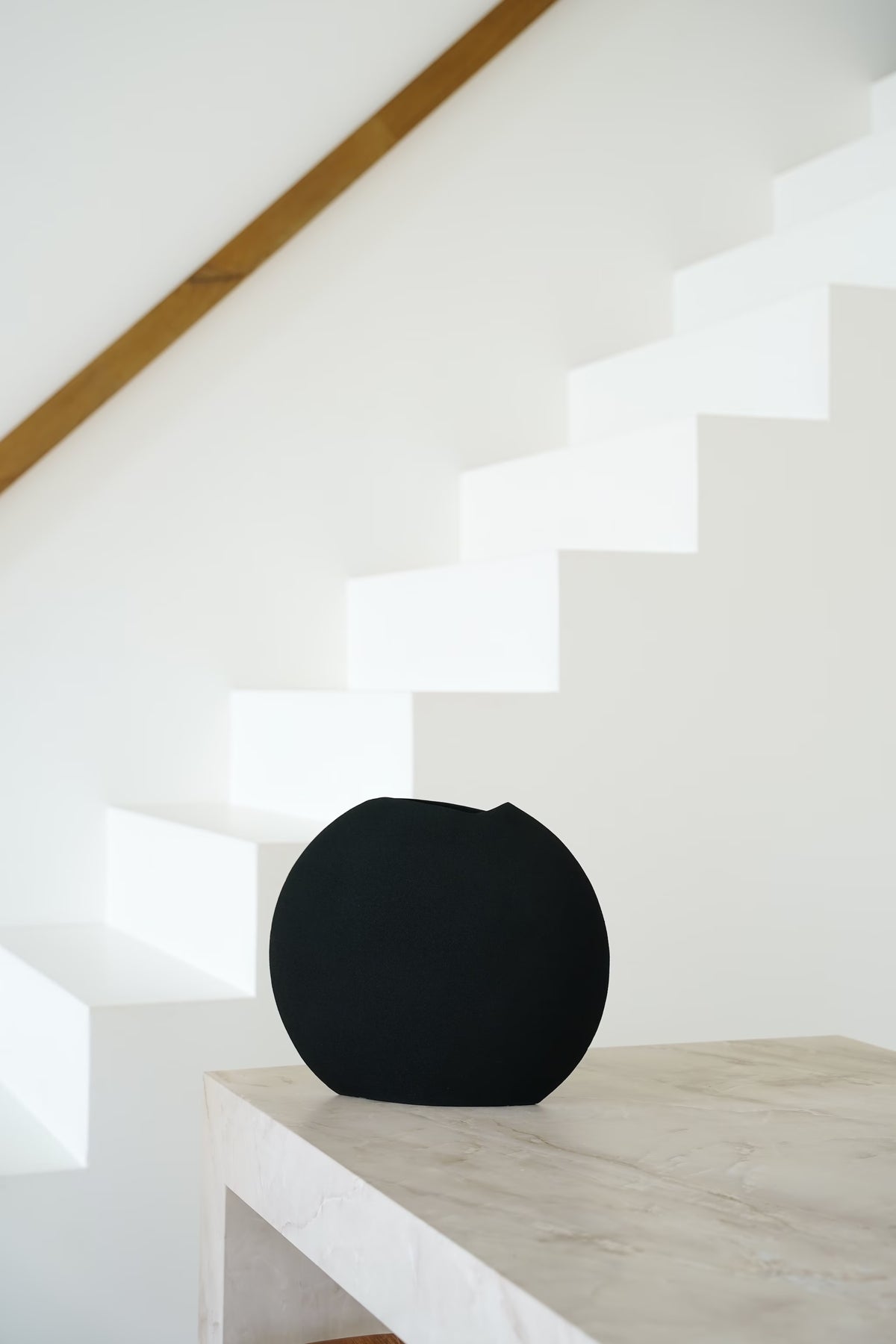 Vooraanzicht van de ronde, matzwarte Vaas Tinta met asymmetrische bovenrand op een lichte stenen sidetable.