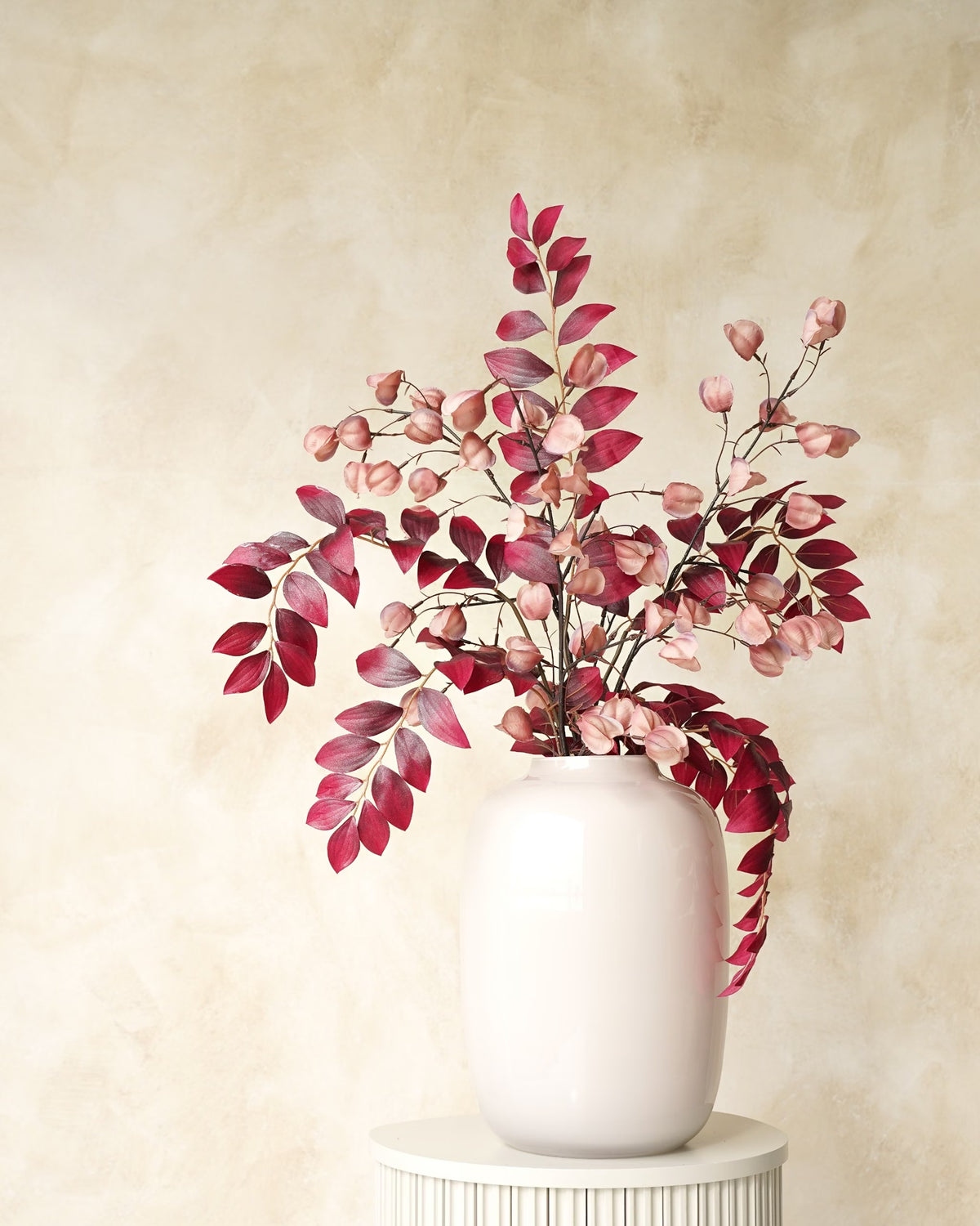 Zijden boeket Amaya van Fan of Flowers met bordeauxrode bladeren en zachtroze lampionnetjes in witte vaas Crema op een ronde sokkel.