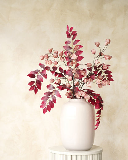 Zijden boeket Amaya van Fan of Flowers met bordeauxrode bladeren en zachtroze lampionnetjes in witte vaas Crema op een ronde sokkel.