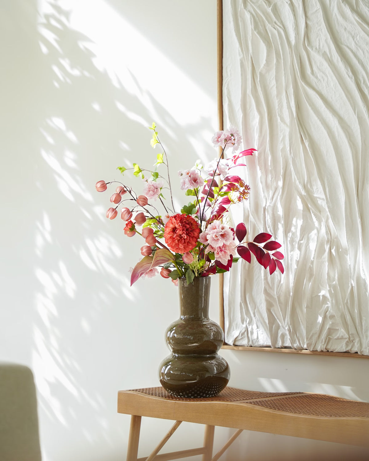 Zijden boeket Carmen van Fan of Flowers met roze bloemen, lampionnetjes en bordeauxrood blad in een bruine glazen vaas op een houten bankje.