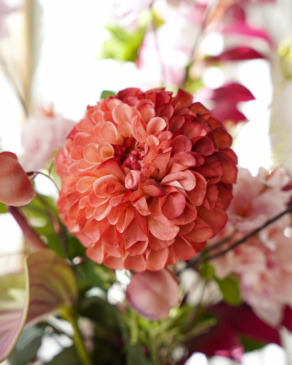 Detail van een volle koraalroze zijden bolbloem in boeket Carmen van Fan of Flowers, omringd door roze lampionnetjes en zachtroze bloesem.