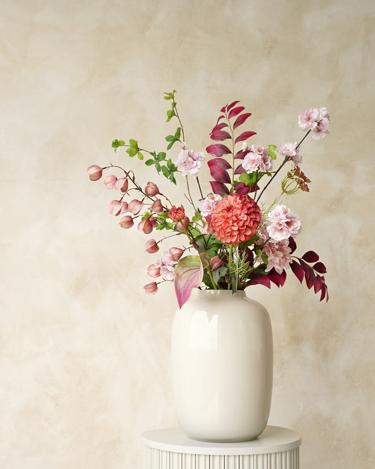 Zijden veldboeket Carmen van Fan of Flowers met zachtroze bloesem, een koraalroze bloem en bordeauxrood blad in witte vaas Crema op een ronde sokkel.