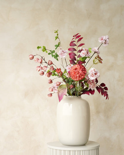 Zijden veldboeket Carmen van Fan of Flowers met zachtroze bloesem, een koraalroze bloem en bordeauxrood blad in witte vaas Crema op een ronde sokkel.