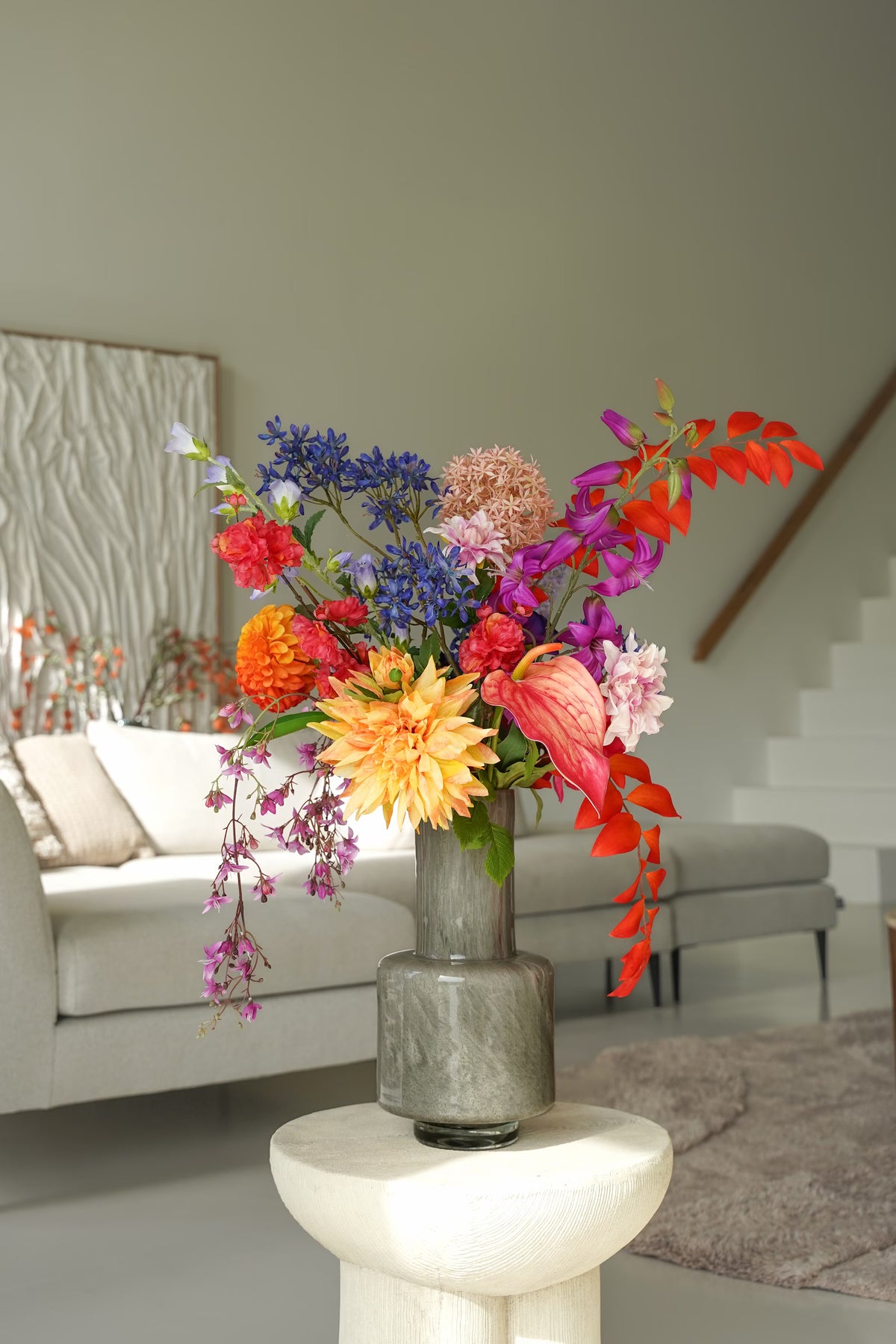 Zijden boeket Chica van Fan of Flowers met oranje, roze, paarse en blauwe bloemen in een grijze vaas met lange hals voor een lichte bank.