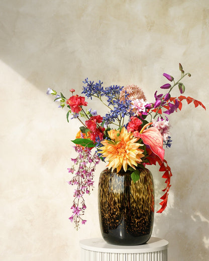 Zijden boeket Chica van Fan of Flowers met een grote geeloranje bloem, roze anthurium en afhangende lila takken in tijgervaas Leo op een ronde sokkel.