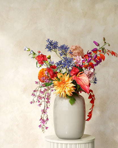 Zijden boeket Chica van Fan of Flowers met kleurrijke bloemen en afhangende lila takken in witte vaas Crema op een ronde sokkel.