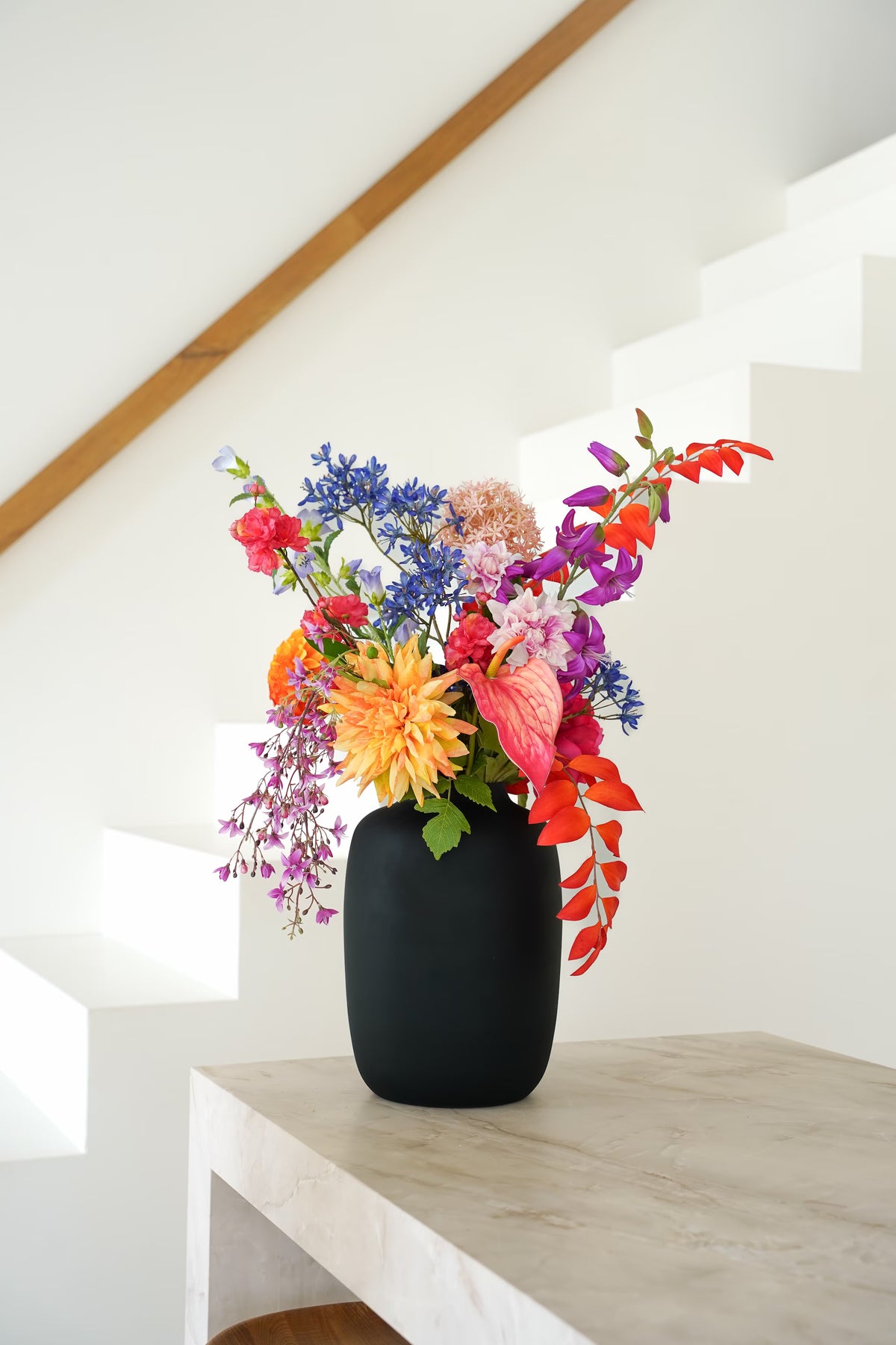 Zijden boeket Chica van Fan of Flowers met oranje, roze, paarse en blauwe bloemen in matzwarte vaas Negro bij een witte trap.