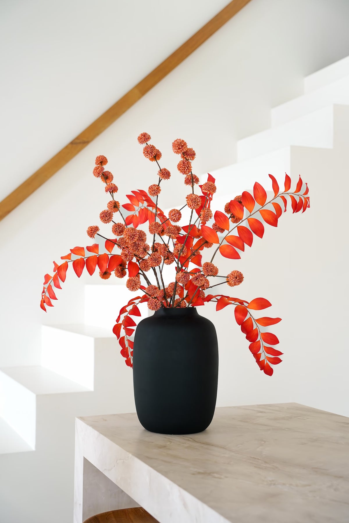 Zijden boeket Fuego van Fan of Flowers met feloranje bladeren en roestkleurige bolbloemen in matzwarte vaas Negro bij een witte trap.