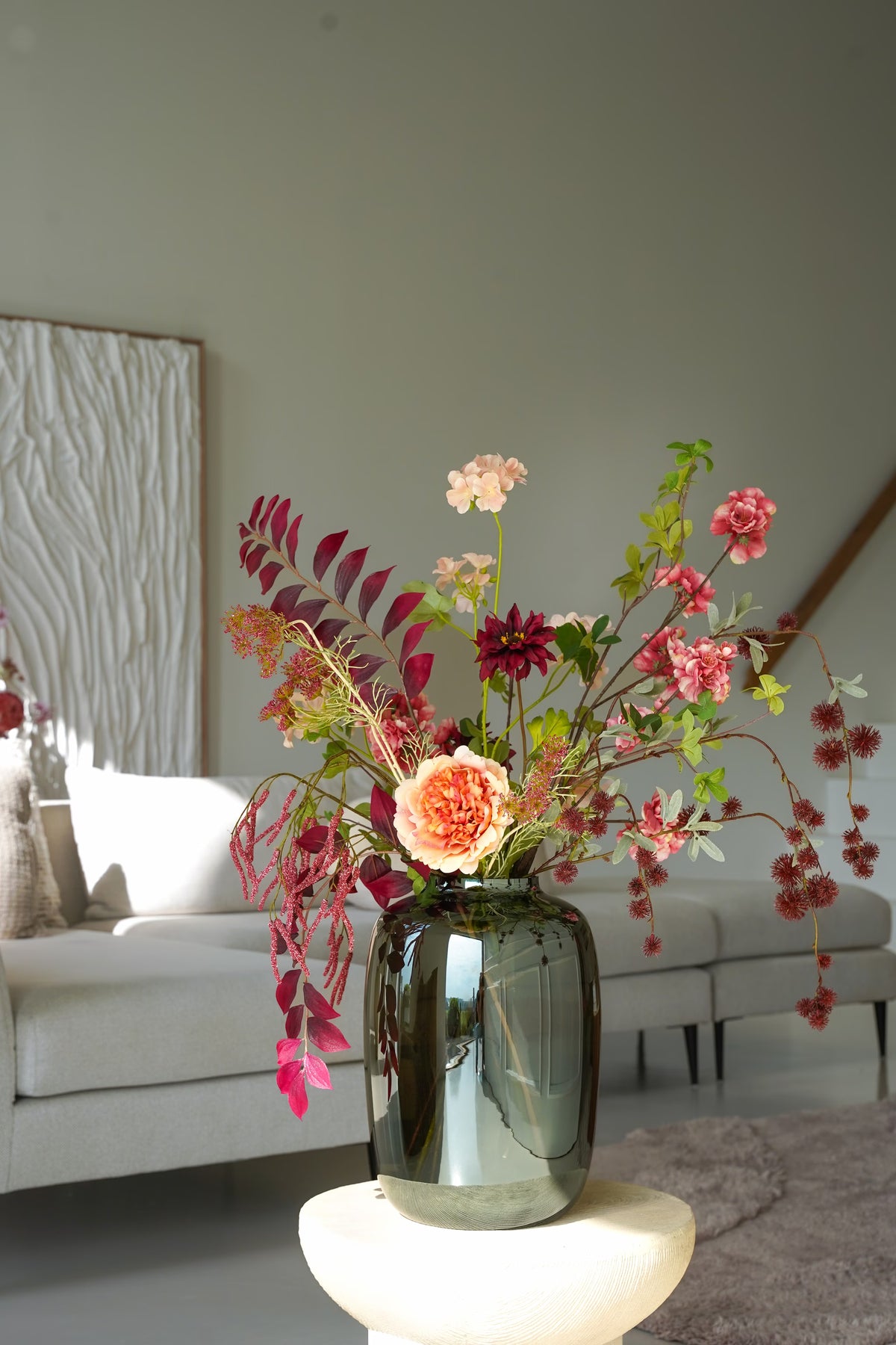 Zijden boeket Lila van Fan of Flowers met bordeauxrode, roze en crèmekleurige bloemen in vaas Glossy op een ronde bijzettafel voor een lichte bank.