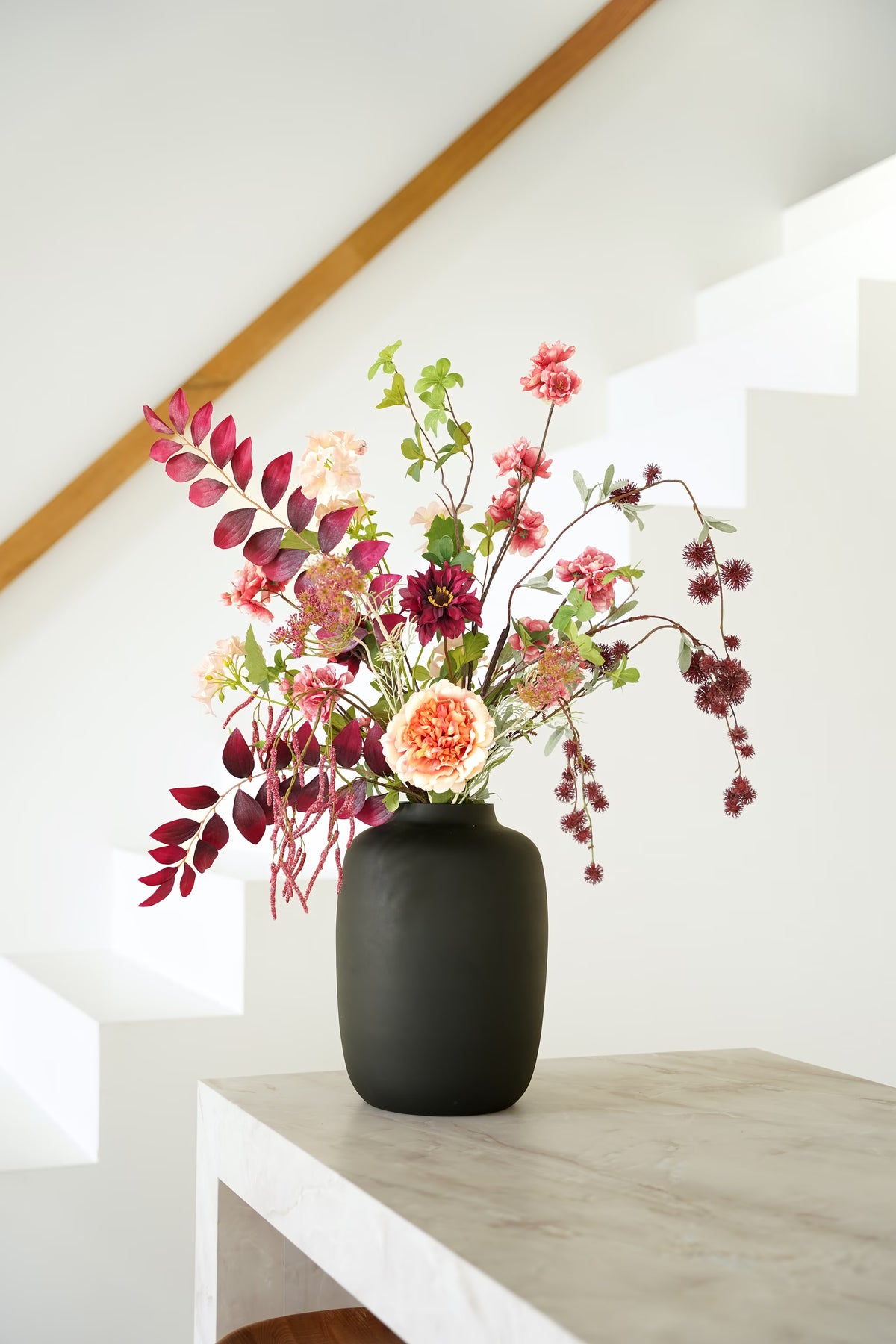 Zijden boeket Lila van Fan of Flowers met bordeauxrood blad, roze en crèmekleurige bloemen in matzwarte vaas Negro bij een witte trap.