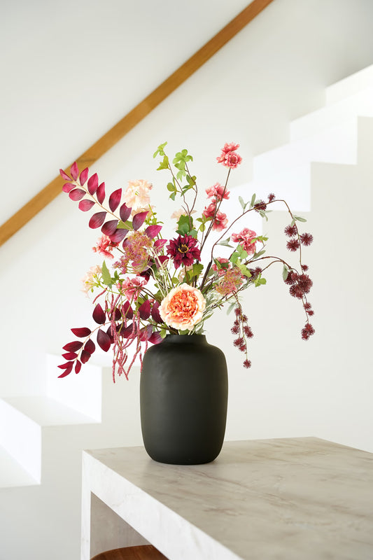 Zijden boeket Lila van Fan of Flowers met bordeauxrood blad, roze en crèmekleurige bloemen in matzwarte vaas Negro bij een witte trap.