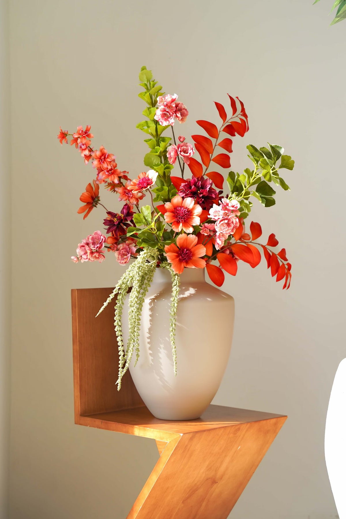 Zijden boeket Palma van Fan of Flowers met oranje en roze bloemen en frisgroene takken in een lichtbeige vaas op een houten stoel.