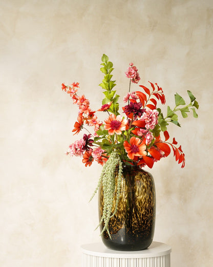 Zijden boeket Palma van Fan of Flowers met oranje en roze bloemen en afhangende groene takken in tijgervaas Leo op een ronde sokkel.