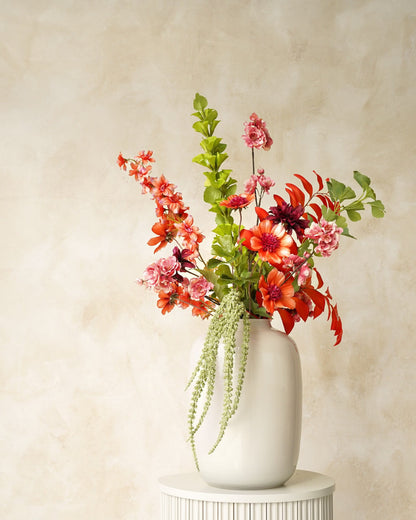 Zijden boeket Palma van Fan of Flowers met oranje bloemen, roze bloesem en groene hangtakken in witte vaas Crema op een ronde sokkel.