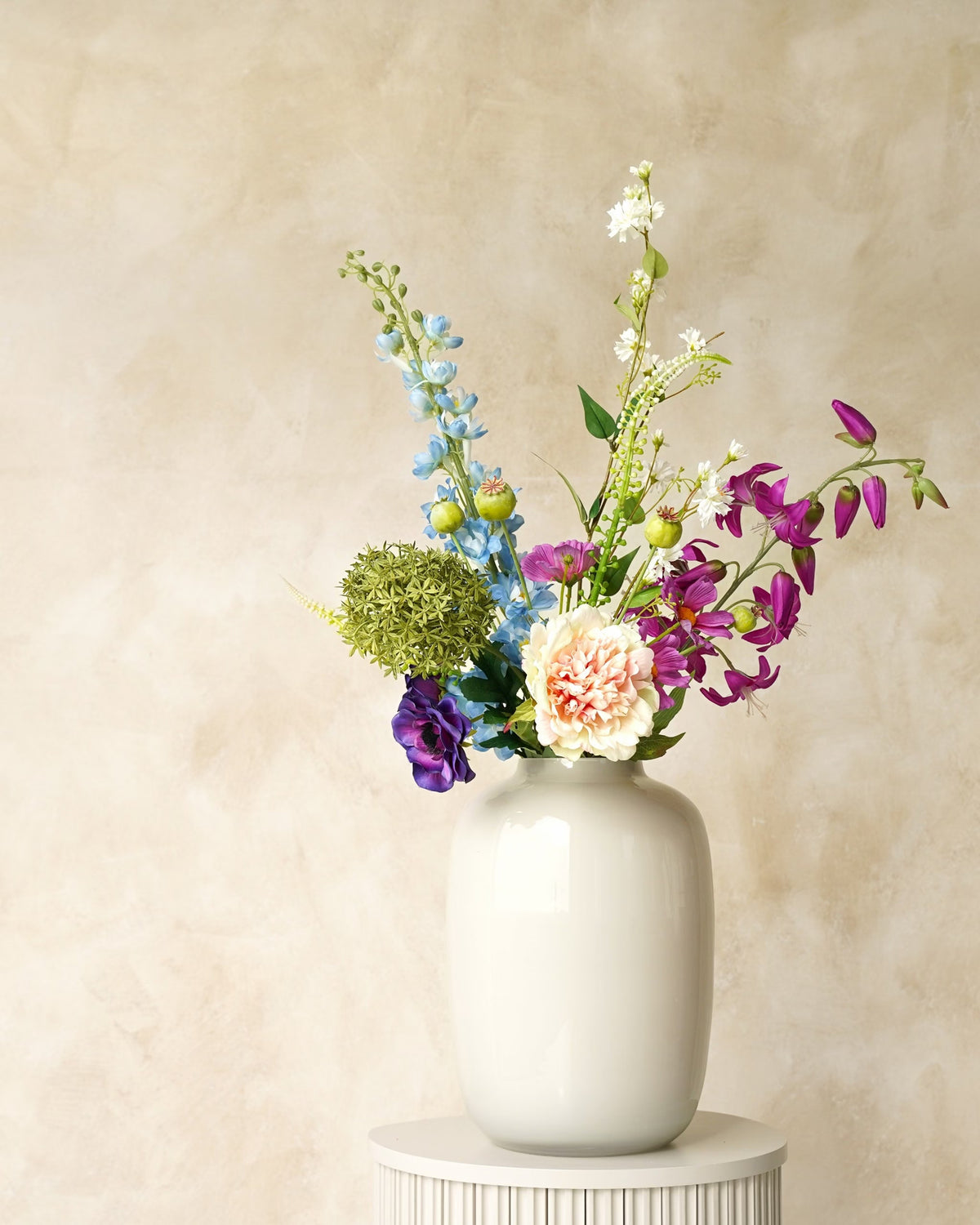 Zijden boeket Rio van Fan of Flowers met lichtblauwe en paarse bloemen, zachte crèmetinten en groen in witte vaas Crema op een ronde sokkel.