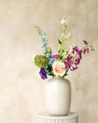 Zijden boeket Rio van Fan of Flowers met lichtblauwe en paarse bloemen, zachte crèmetinten en groen in witte vaas Crema op een ronde sokkel.