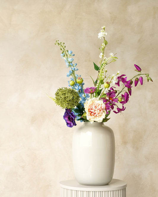 Zijden boeket Rio van Fan of Flowers met lichtblauwe en paarse bloemen, zachte crèmetinten en groen in witte vaas Crema op een ronde sokkel.