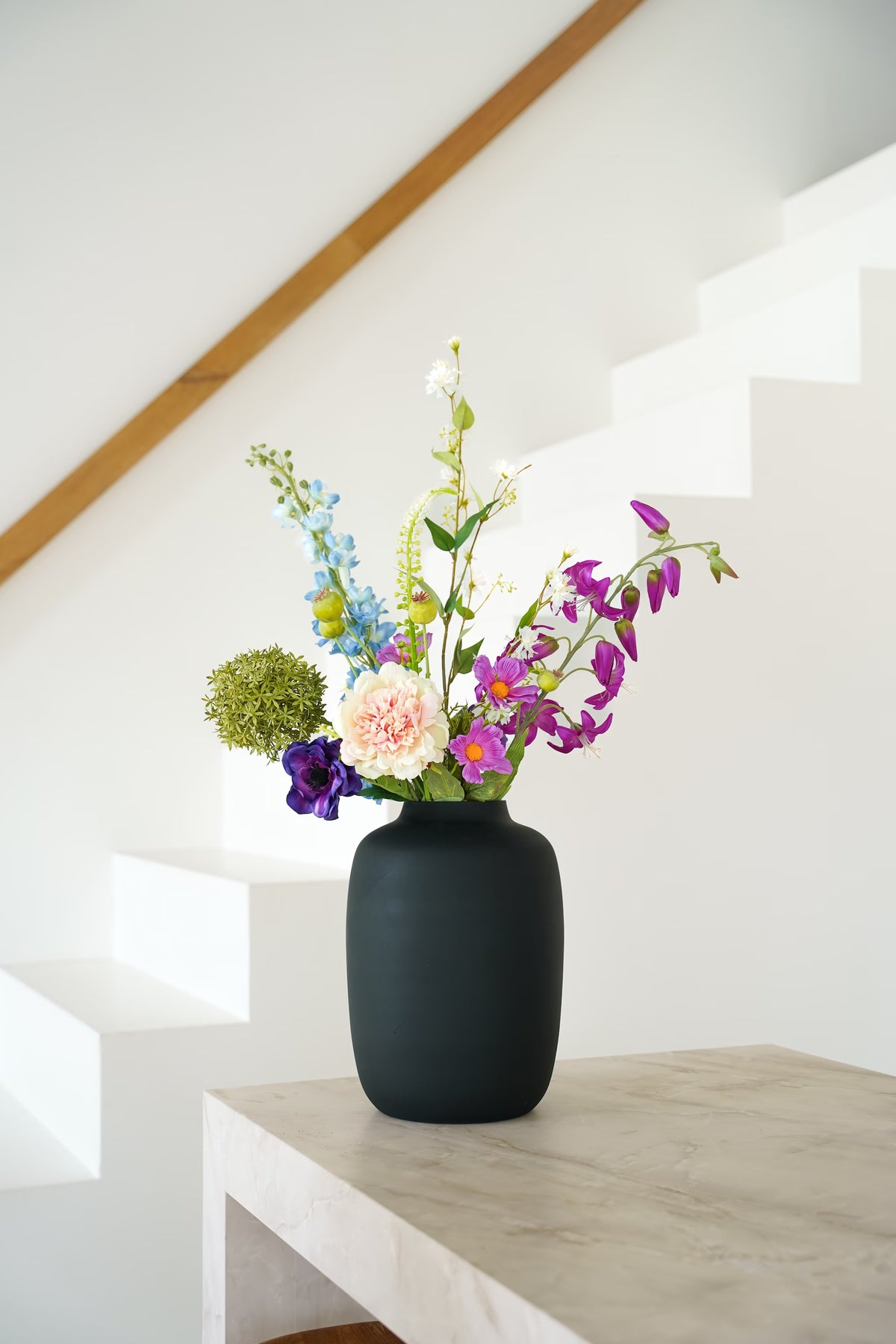 Zijden boeket Rio van Fan of Flowers met lichtblauwe, paarse en crèmekleurige bloemen in matzwarte vaas Negro bij een witte trap.