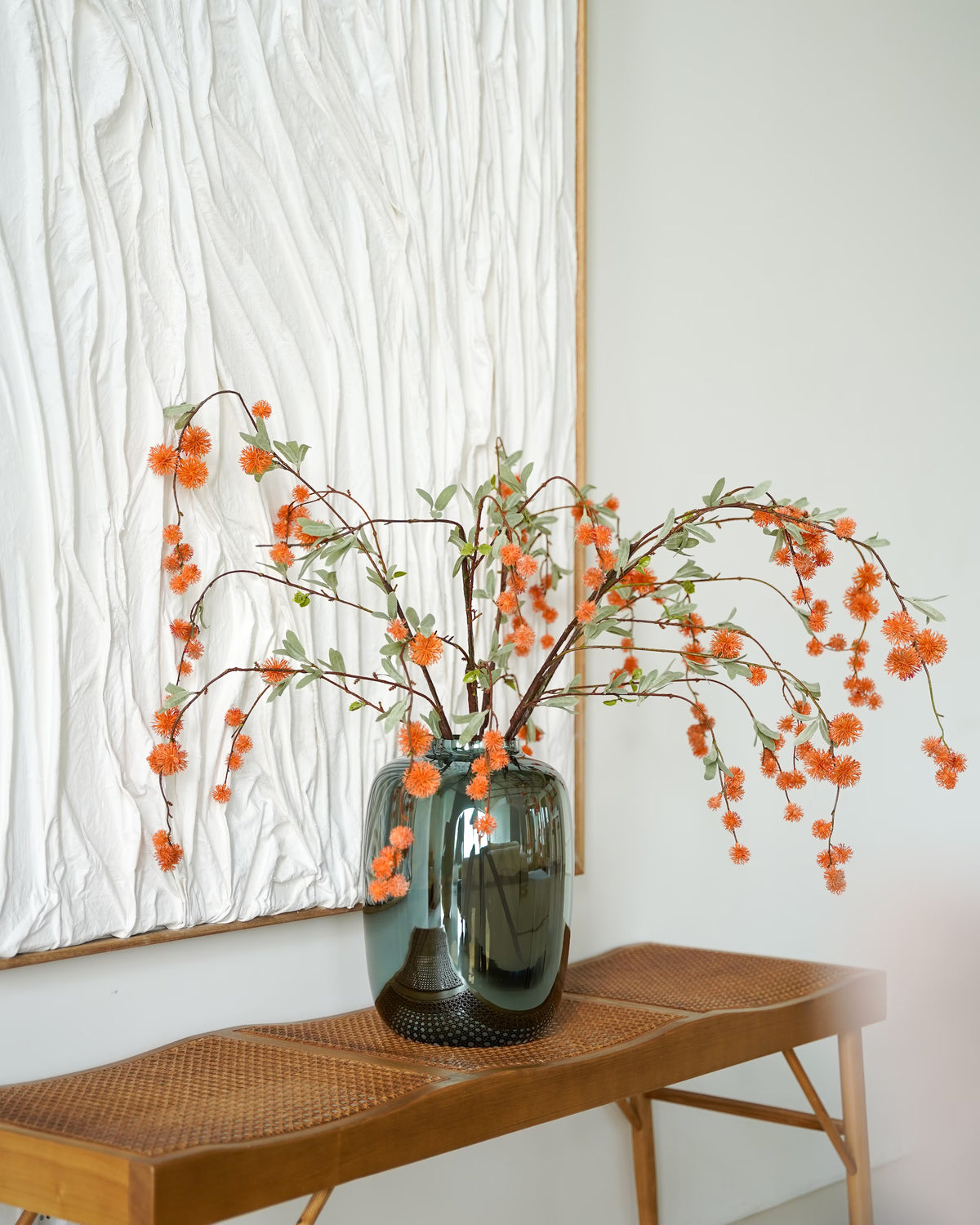 Oranje zijden bolletjestakken van Fan of Flowers in een glanzende glazen vaas op een houten bankje voor een wit wandkunstwerk.