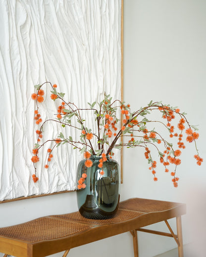 Oranje zijden bolletjestakken van Fan of Flowers in een glanzende glazen vaas op een houten bankje voor een wit wandkunstwerk.