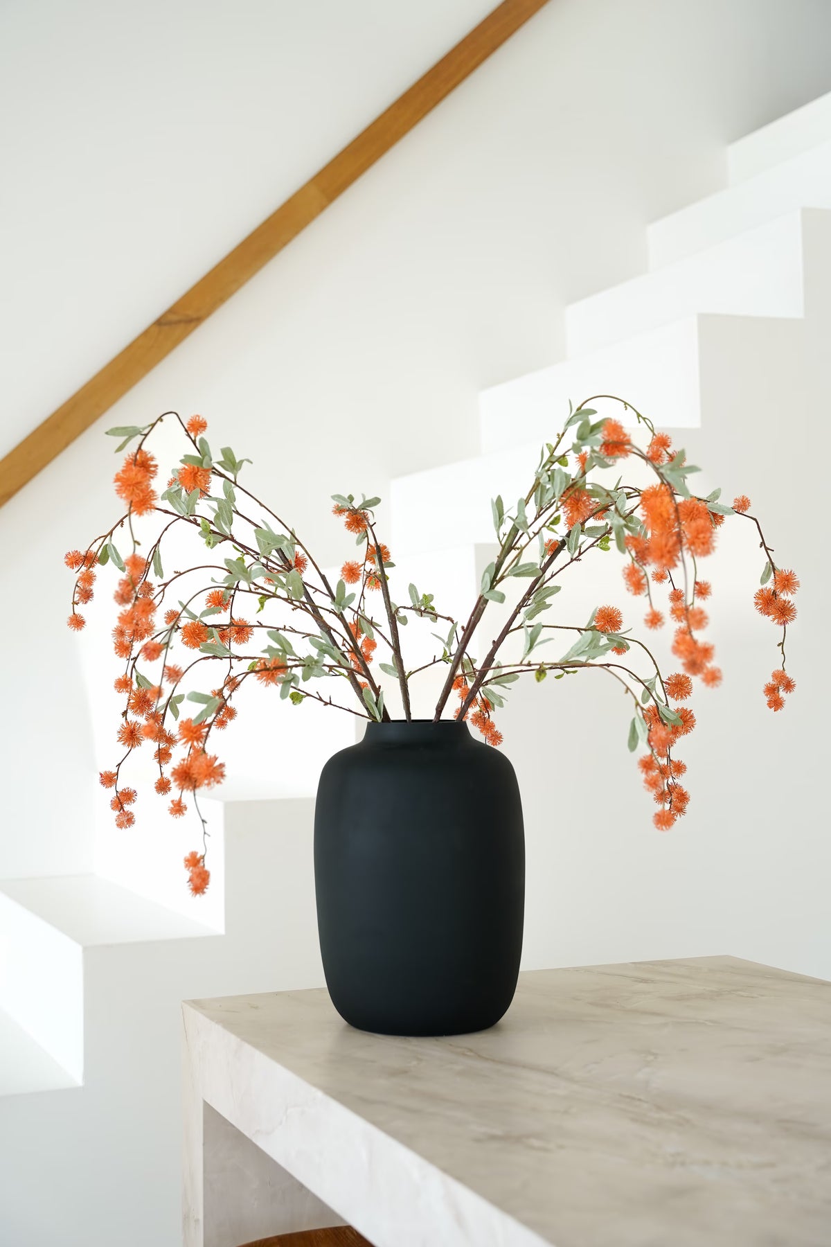 Oranje zijden bolletjestakken van Fan of Flowers met grijsgroene blaadjes in matzwarte vaas Negro bij een witte trap.